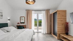 A modern hotel room with a large bed, a desk, a chair, a wooden wardrobe, and a view of the outdoors through a sliding door.