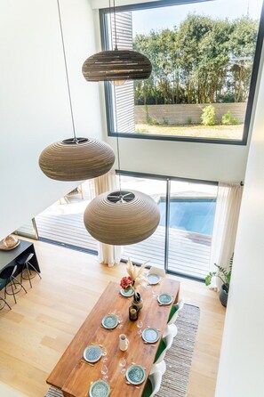 A dining area with a wooden table set for four, hanging pendant lights, and a view of a swimming pool through large windows.