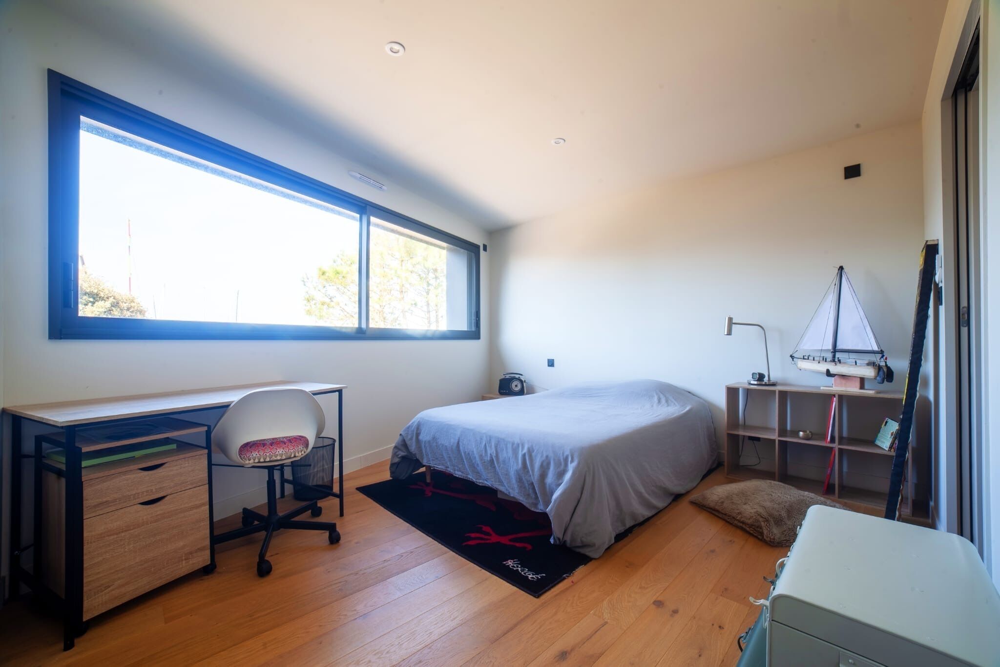 Een slaapkamer met een bureau, stoel, bed en een groot raam.