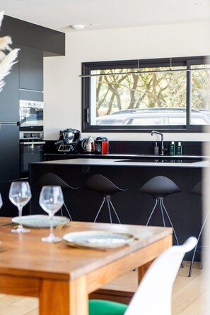 A modern kitchen with a large window, a dining area with a wooden table and chairs, and a view of trees outside.