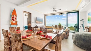 A dining area with a wooden table set for six, surrounded by wicker chairs, a large window with a view of the ocean, and a ceiling fan.