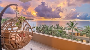 A balcony with a wicker chair, a view of palm trees, and a sunset over the ocean.