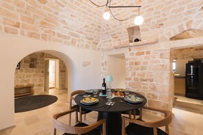 Une pièce aux murs de pierre, avec une table ronde dressée pour le repas, un banc et une porte-fenêtre donnant sur une autre pièce.