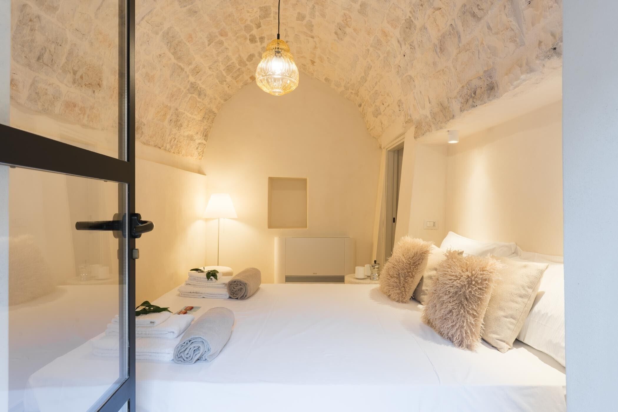 A hotel room with a stone ceiling, a bed with white linens, a bedside table with a lamp, and a mirror.