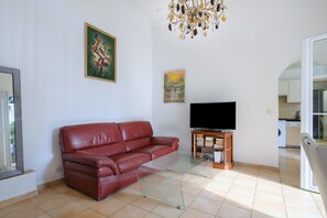 A living room with a red leather sofa, a flat-screen TV on a wooden stand, a glass coffee table, a chandelier, and a painting on the wall.