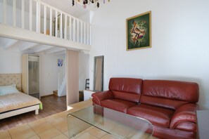 A room with a glass coffee table, a red leather sofa, a bed with a light-colored headboard, and a staircase leading to an upper level.