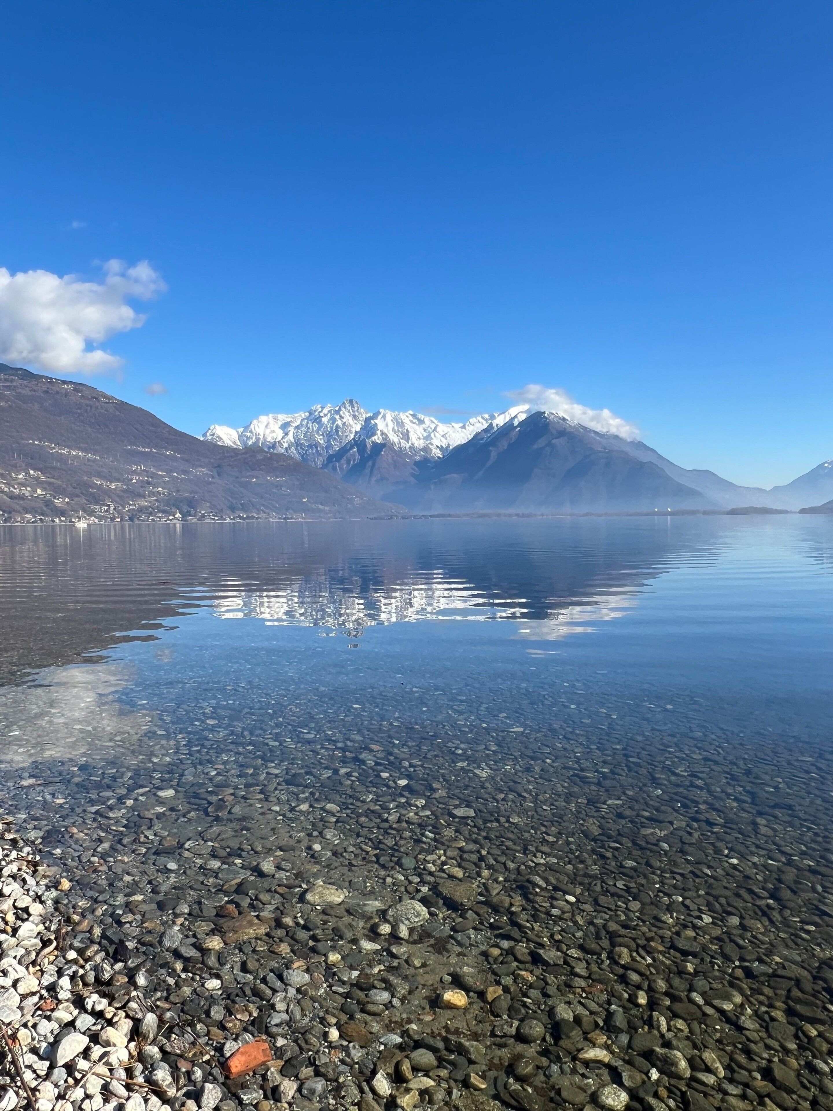 A clear lake with a rocky shoreline and snow-capped mountains in the background.