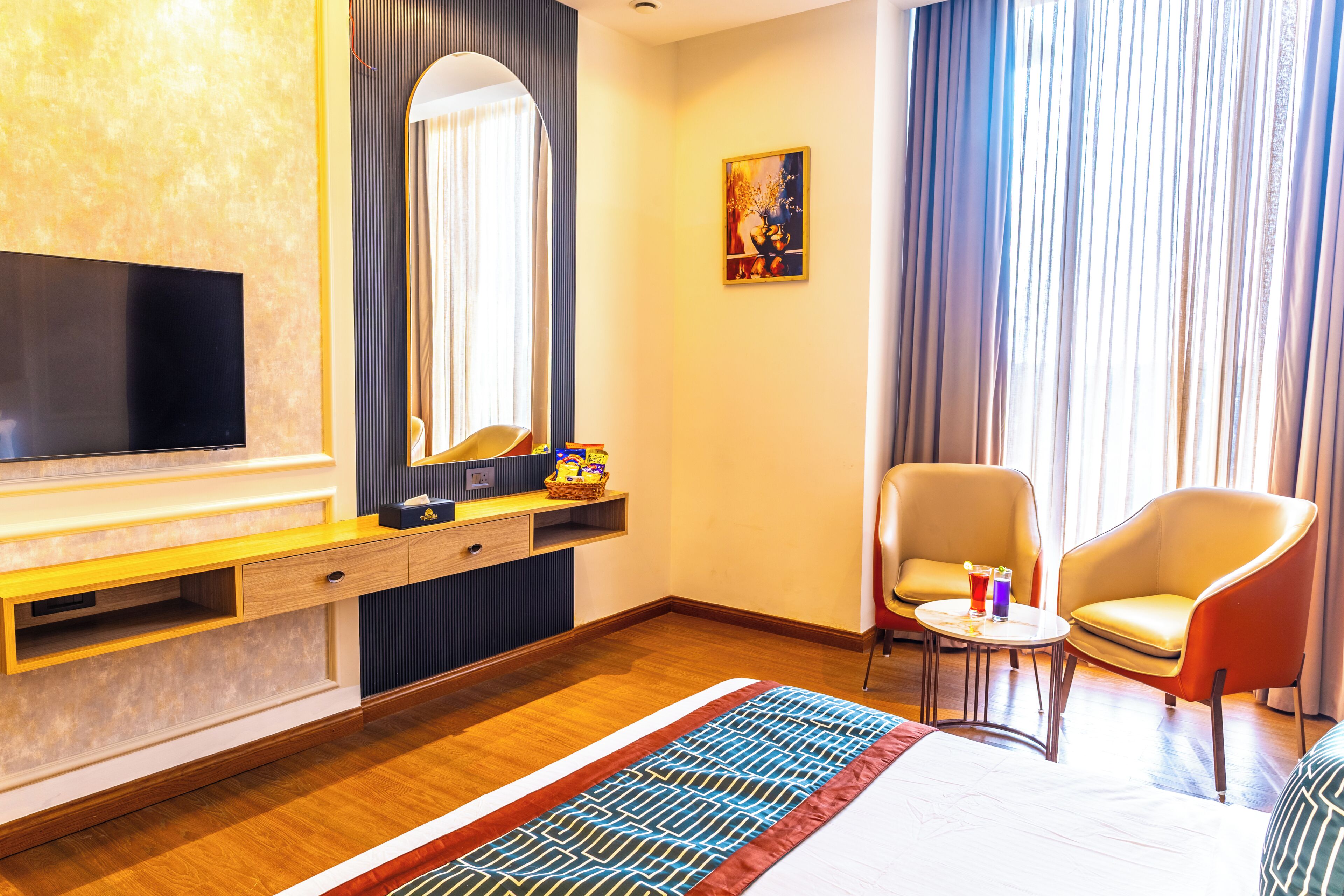 A hotel room with a bed, a TV, a desk with drawers, a chair, a small table with drinks, and a painting on the wall.
