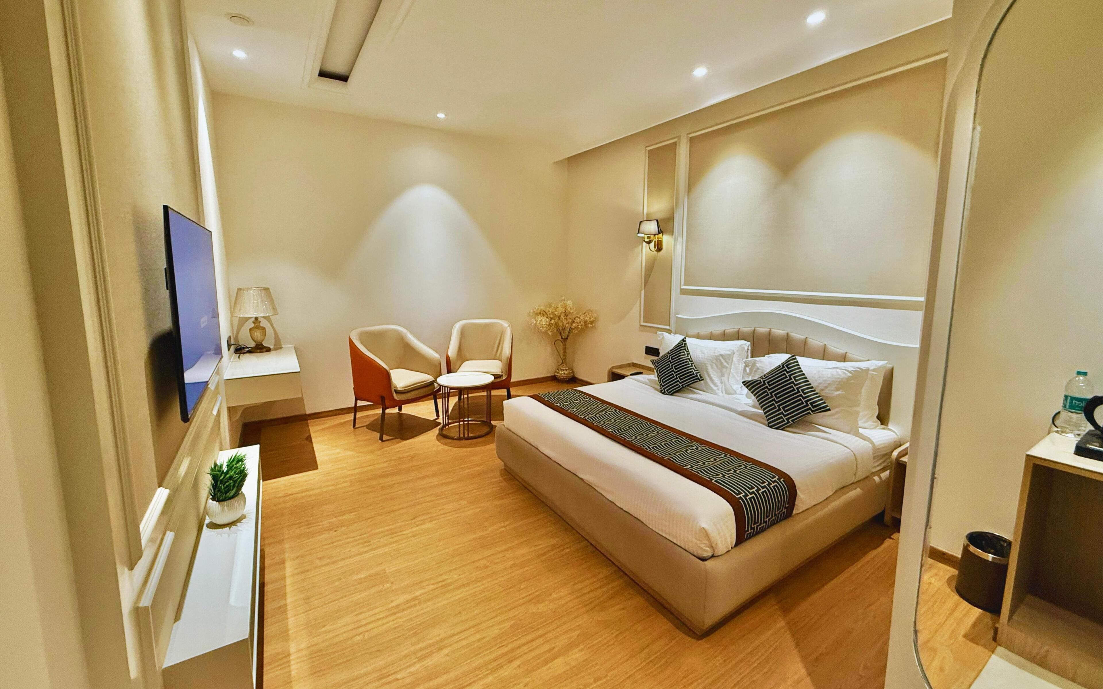 A modern hotel room with a large bed, a desk with a lamp, two chairs, a small table, and a TV.