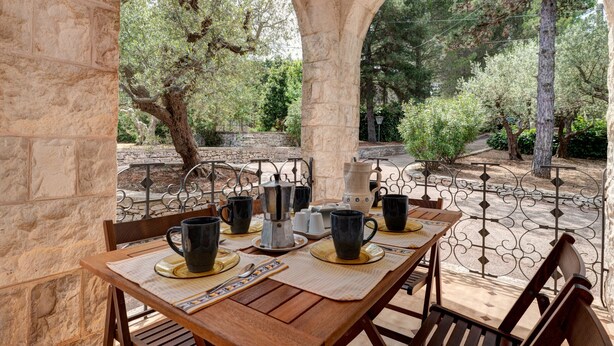 Un área de comedor al aire libre con pared de piedra, una mesa de madera con tazas y platos negros, y vistas a árboles y una pared de piedra al fondo.
