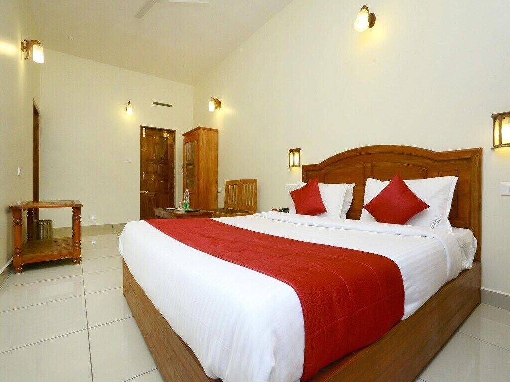 A hotel room with a large bed, wooden headboard, white bedding, and red pillows. There is a wooden nightstand with a lamp, a wooden wardrobe, and a door in the background.