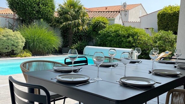 Un espace repas extérieur avec vue sur la piscine, comprenant une table dressée pour quatre personnes avec des chaises, une pergola au-dessus et une piscine en arrière-plan.