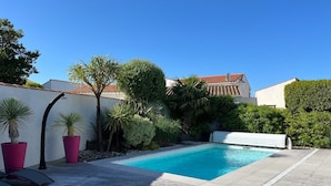 Une piscine dotée d’une terrasse en bois, entourée de plantes en pot et bordée d’un mur blanc.