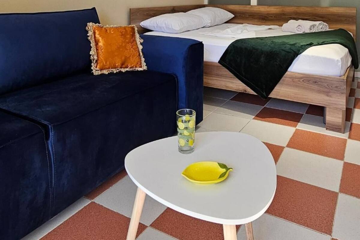 A room with a dark blue sofa, a white table with a yellow bowl, a bed with a green blanket, and a tall glass of water.