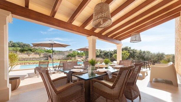Un área de comedor al aire libre techada, con sillas de mimbre y una mesa de vidrio, con vistas a una piscina y un paisaje exuberante.