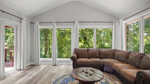A spacious living room with a large sectional sofa, a round coffee table, and large windows offering a view of greenery outside.