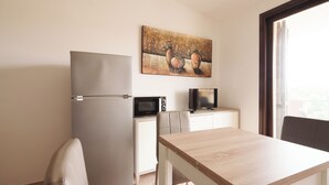 Una kitchenette compacta con refrigerador, microondas y un televisor pequeño sobre un mueble blanco. Hay una mesa de madera y una silla, y un cuadro cuelga en la pared.