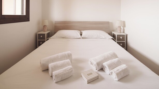 Una cama bien hecha con sábanas blancas y toallas enrolladas, dos mesitas de noche con lámparas, y una ventana a la izquierda.