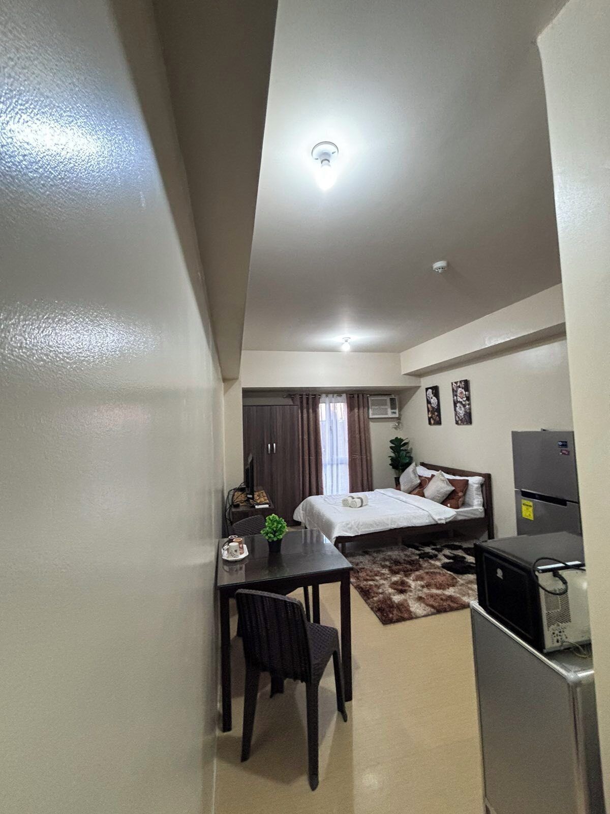 A compact hotel room with a bed, a desk, a chair, a small table, a TV, and a refrigerator.