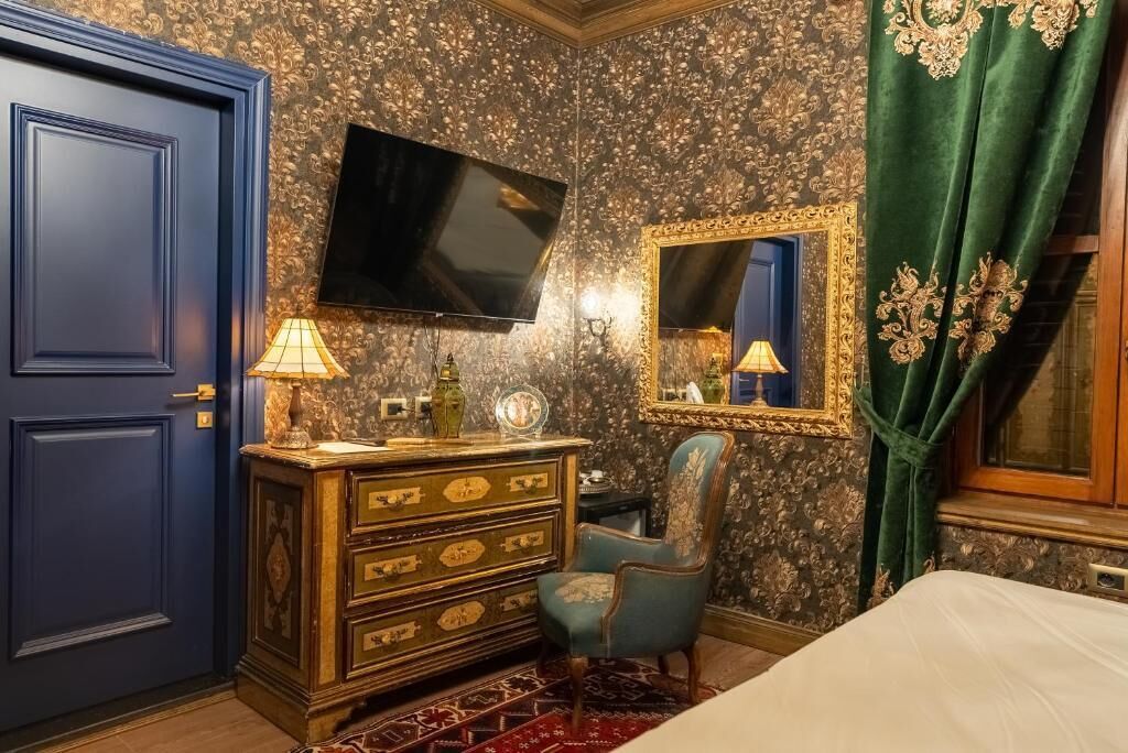 A room with a blue door, a dresser with drawers, a chair, a bed, a television mounted on the wall, and a mirror with a gold frame.