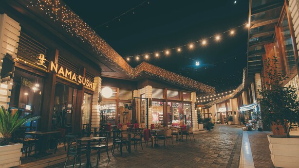 Un espace de restauration en plein air avec des tables et des chaises, un restaurant de sushis et des guirlandes lumineuses.