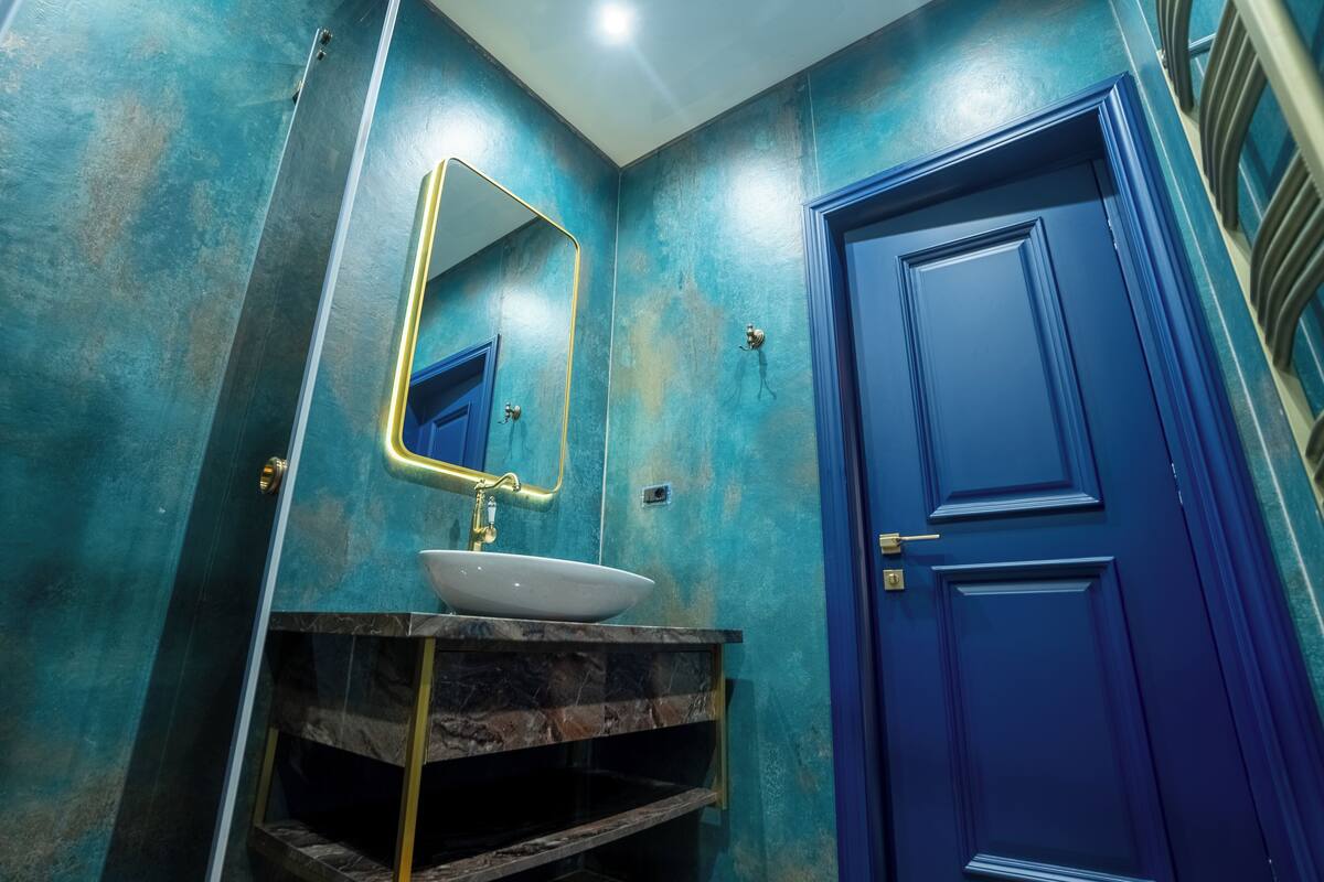Une salle de bain avec une porte bleue, un lavabo blanc avec un robinet doré et un miroir avec un cadre doré.