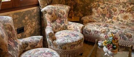 A room with floral-patterned armchairs and a sofa, a glass coffee table with a floral arrangement, and a window with wooden frames.