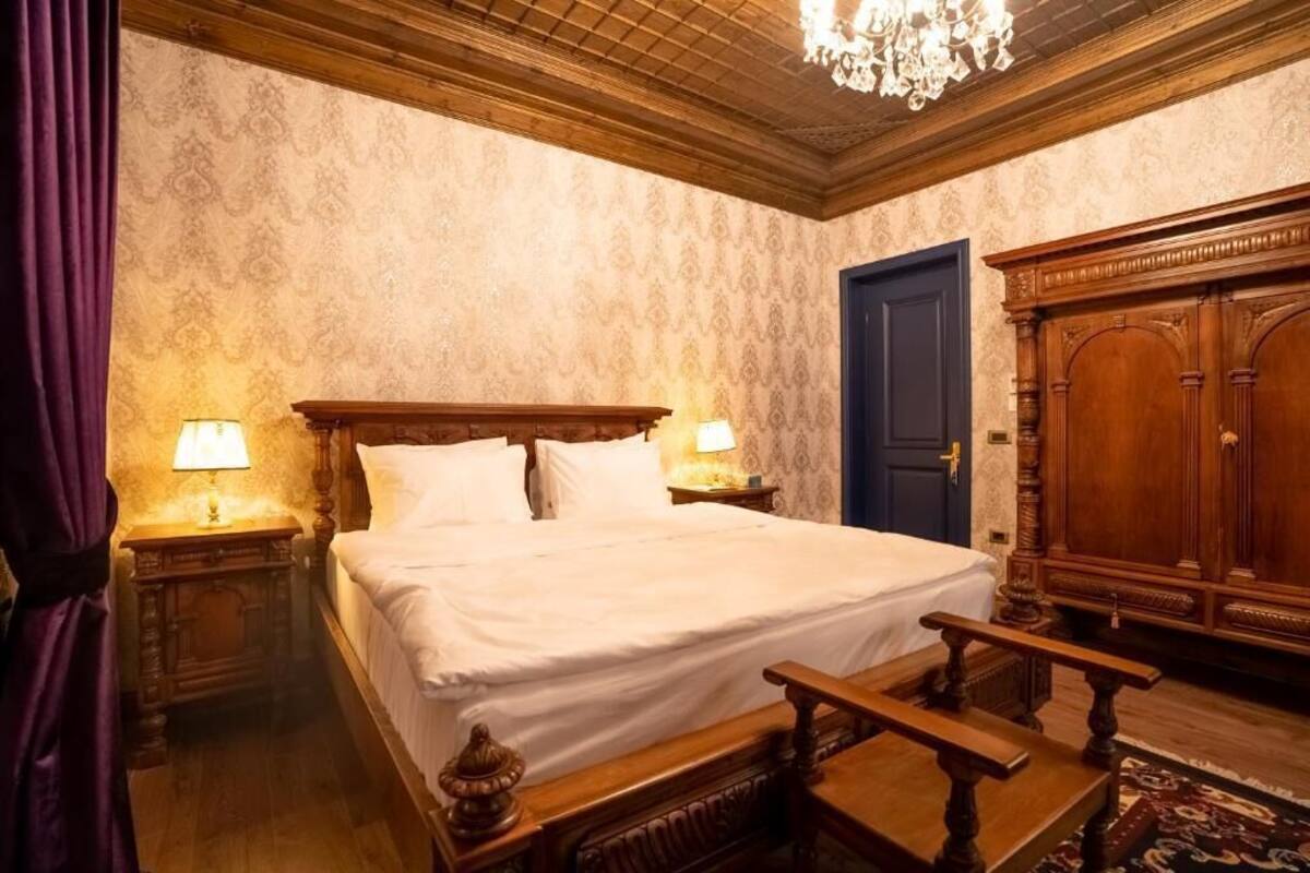 Une chambre d’hôtel avec un grand lit, une tête de lit en bois, deux lampes de chevet, une armoire en bois et un lustre.