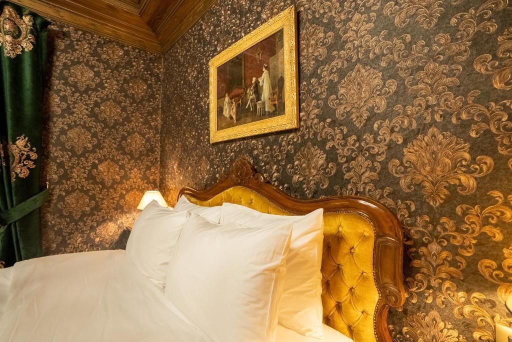 A bed with white linens and a tufted headboard, set against a wall with ornate wallpaper and a framed painting above.