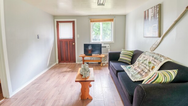 Een woonkamer met een houten vloer, een donkerbank met gestreepte kussens, een houten salontafel, een televisie op een meubelstuk, en een raam met zonwering.
