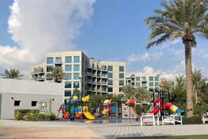 A playground with various play structures, surrounded by palm trees and a building with multiple balconies.