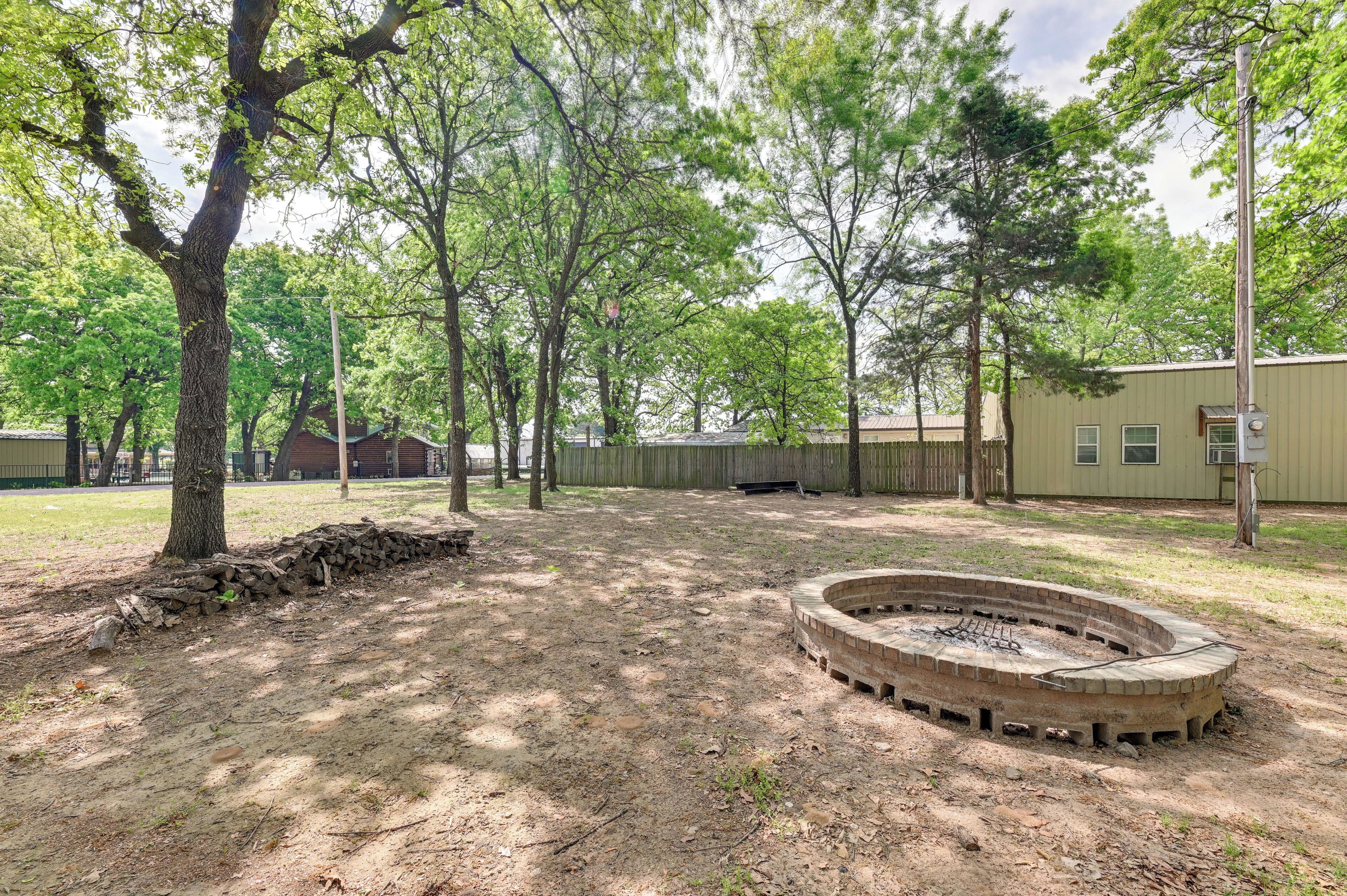 A fenced outdoor area with a circular stone fire pit, surrounded by trees and a building in the background.