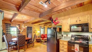 L’intérieur d’une cabane en rondins comprend un coin cuisine équipé d’un réfrigérateur, d’un four à micro-ondes et d’une cuisinière. On y trouve des chaises et une table en bois, ainsi qu’une fenêtre avec des rideaux.