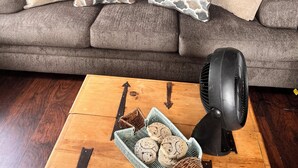 Un salon avec un canapé gris, des coussins à motifs, une table basse en bois agrémentée d’objets décoratifs et un ventilateur.
