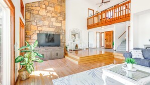 A spacious living room with a stone fireplace, a flat-screen TV, wooden flooring, a balcony, and a staircase. - Swim & Boat Docks: Lakefront Gem in Bella Vista! (Bella Vista)