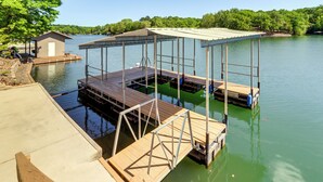 A dock with a covered seating area and a small building, likely a restroom, by a calm lake with trees in the background. - Swim & Boat Docks: Lakefront Gem in Bella Vista! (Bella Vista)
