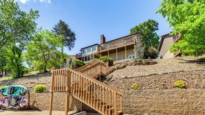 A multi-story house with a wooden staircase leading to a balcony, surrounded by trees and a stone retaining wall. - Swim & Boat Docks: Lakefront Gem in Bella Vista! (Bella Vista)