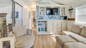 Ein wohnzimmer im Strandhausstil mit Fernseher, Regalen mit Dekoration und zwei beigefarbenen Sofas.