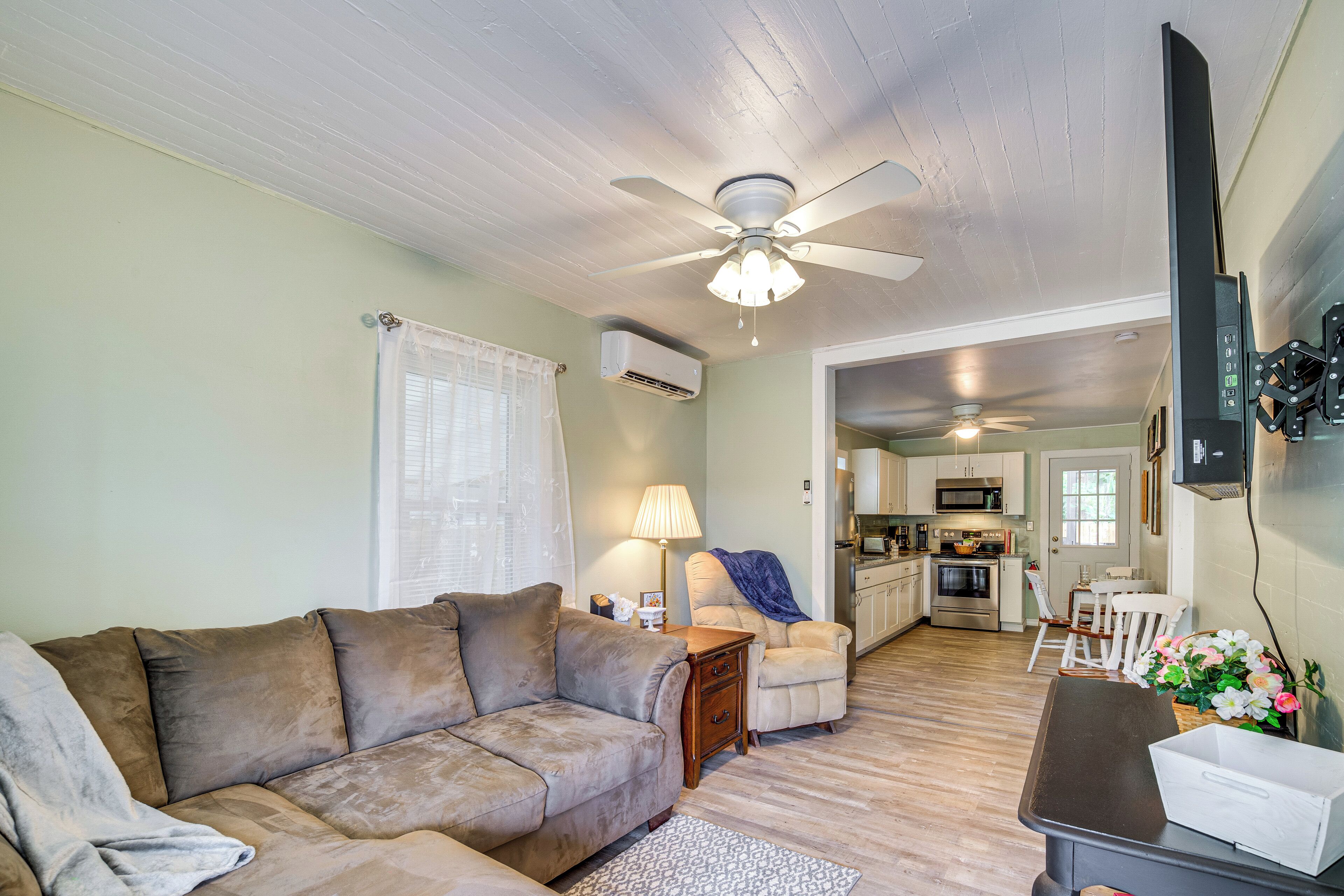 A living room with a sofa, a coffee table, and a ceiling fan. There is a kitchen in the background with a microwave, oven, and sink.