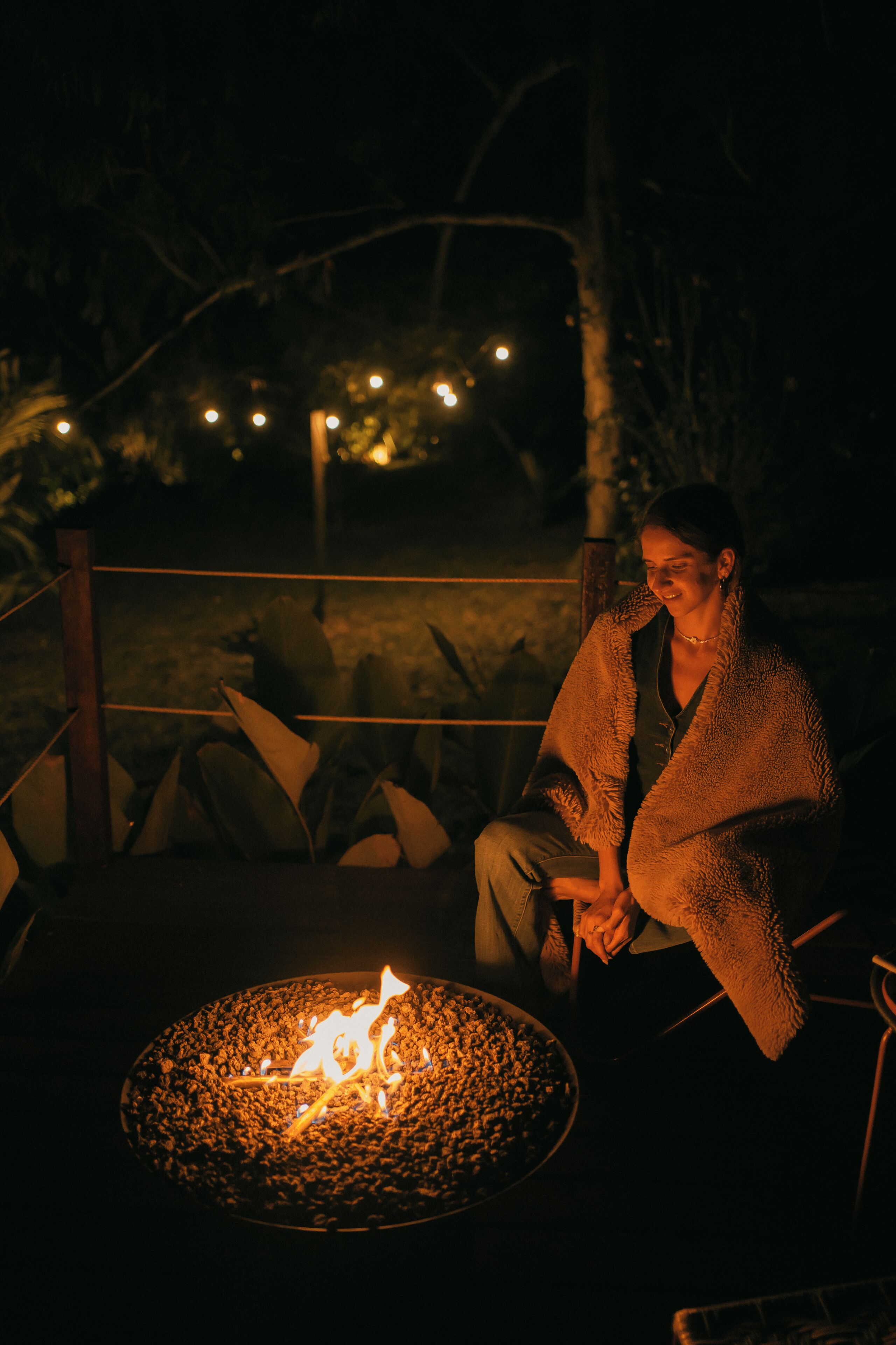 Uma pessoa sentada perto de uma fogueira em área externa com iluminação fraca e luzes de corda ao fundo.