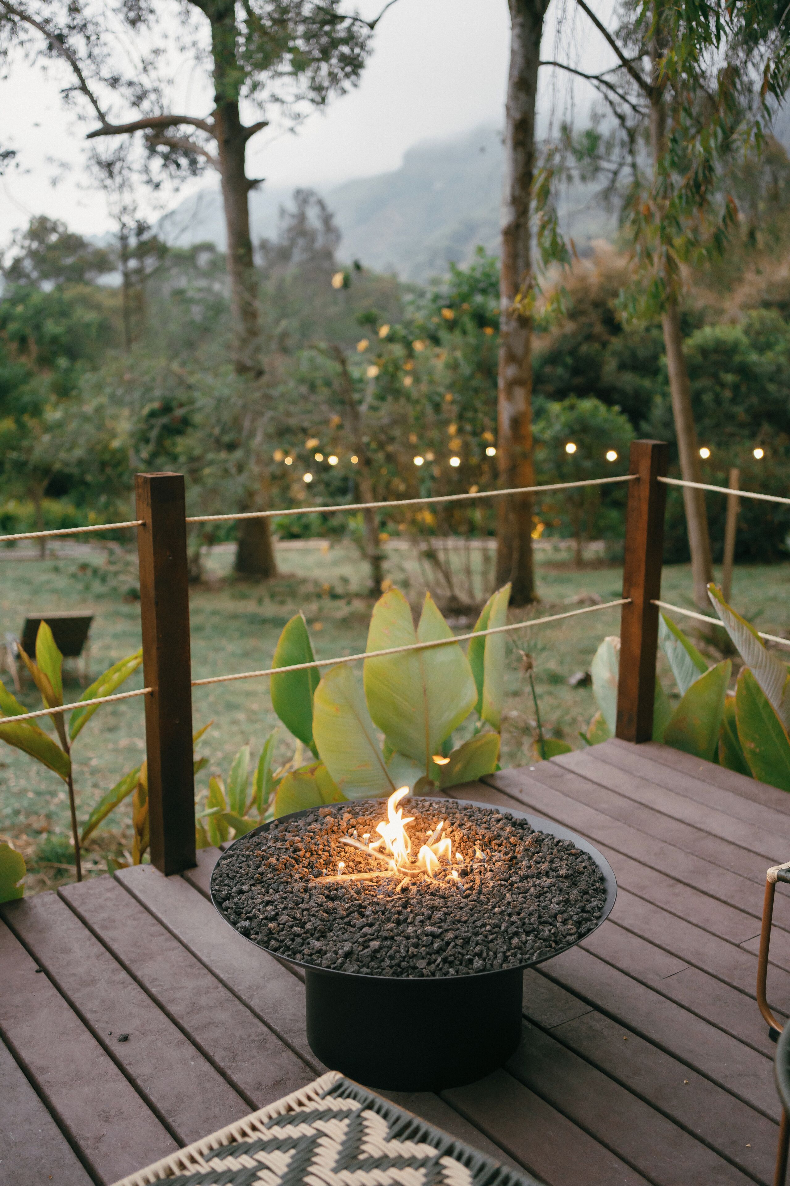 A round outdoor fire pit with a visible flame, surrounded by a wooden deck and greenery.