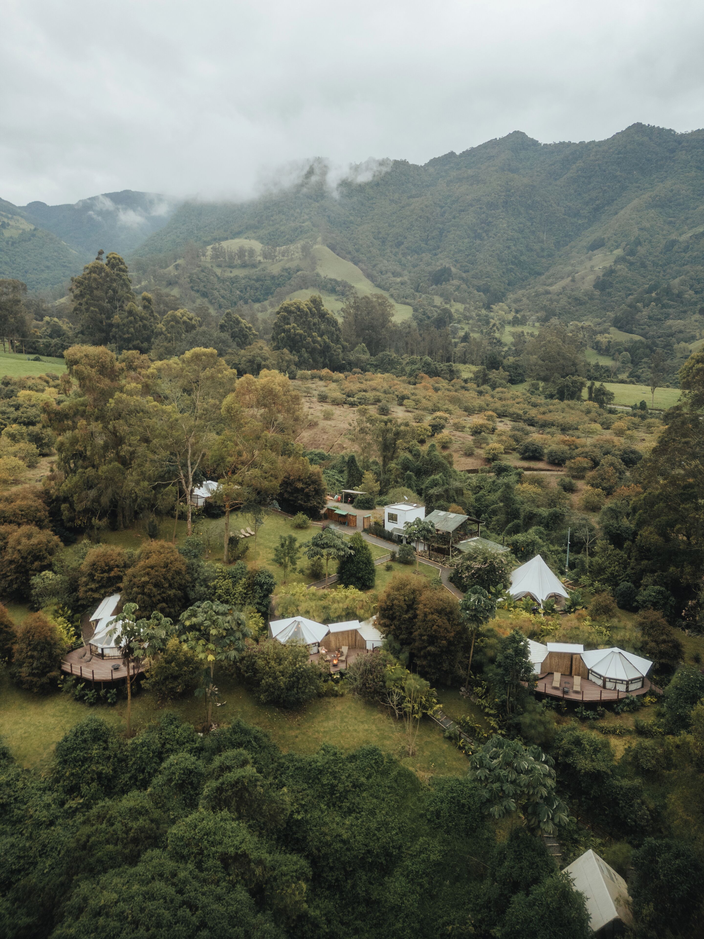 An aerial view of a remote eco-lodge with several dome-shaped cabins surrounded by dense forest and hills.