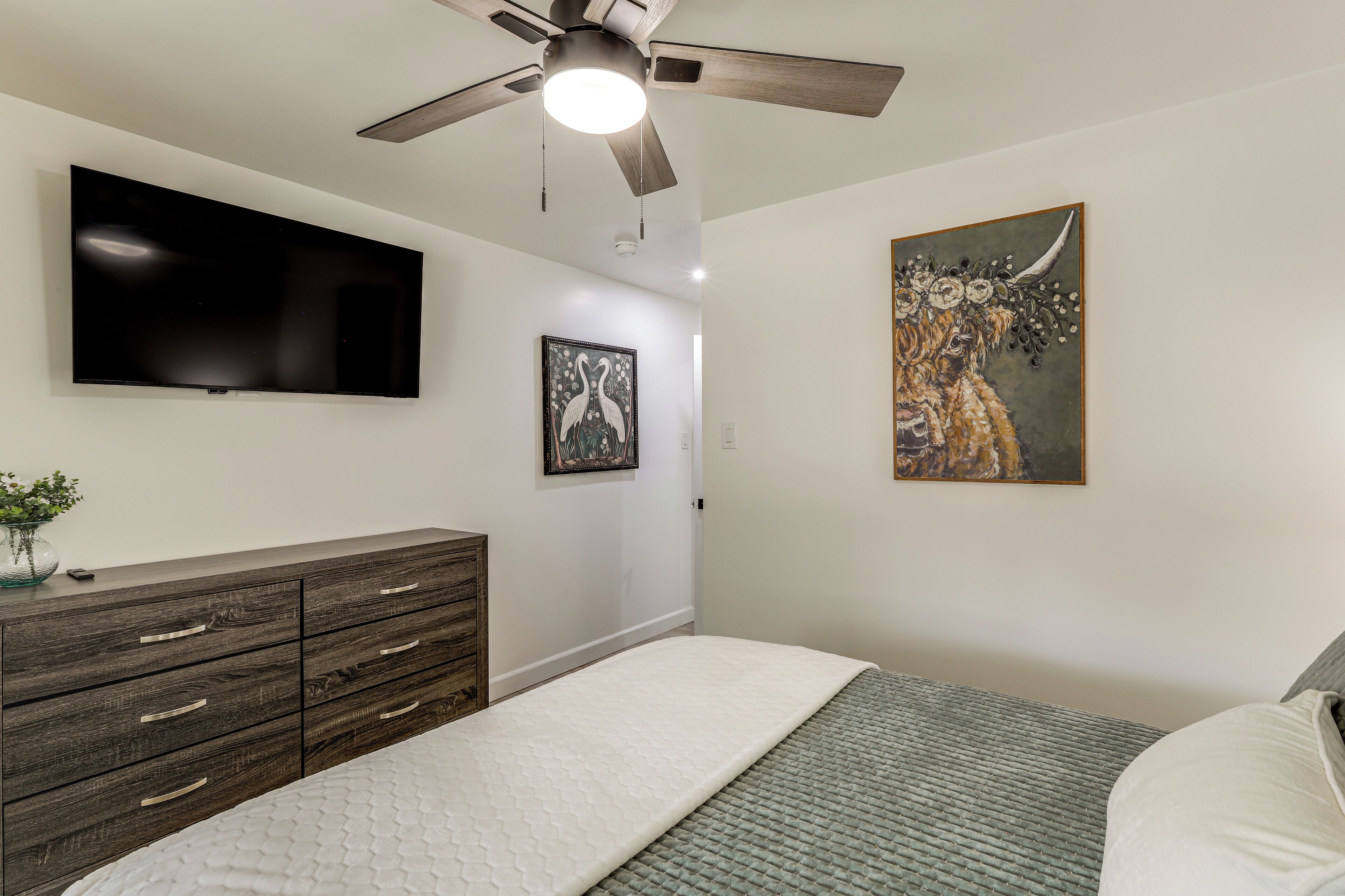 A bedroom with a bed, a dresser, a ceiling fan, a TV, and two framed artworks.