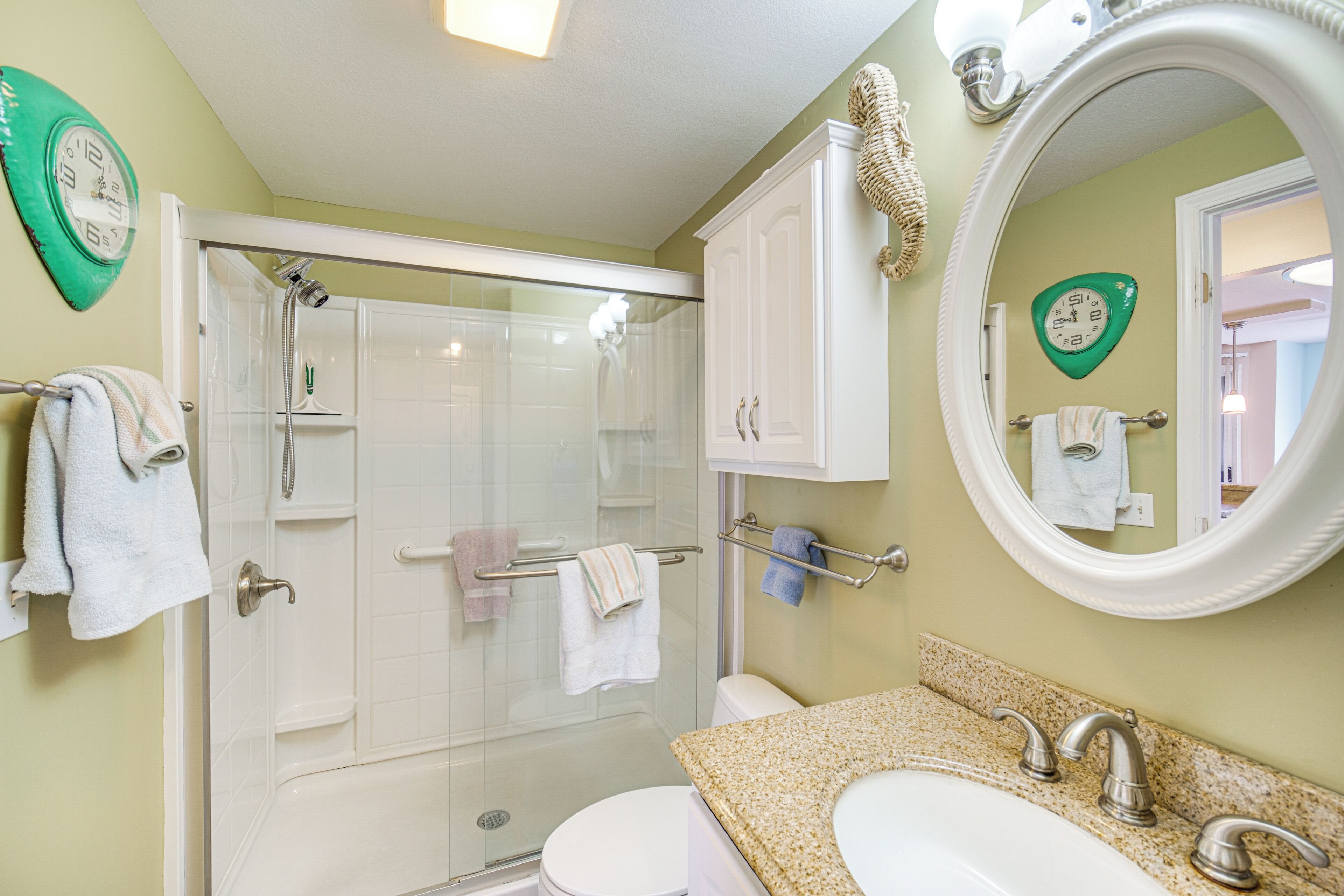 A bathroom with a glass-enclosed shower, a white toilet, a sink with a granite countertop, and a mirror with a decorative frame.