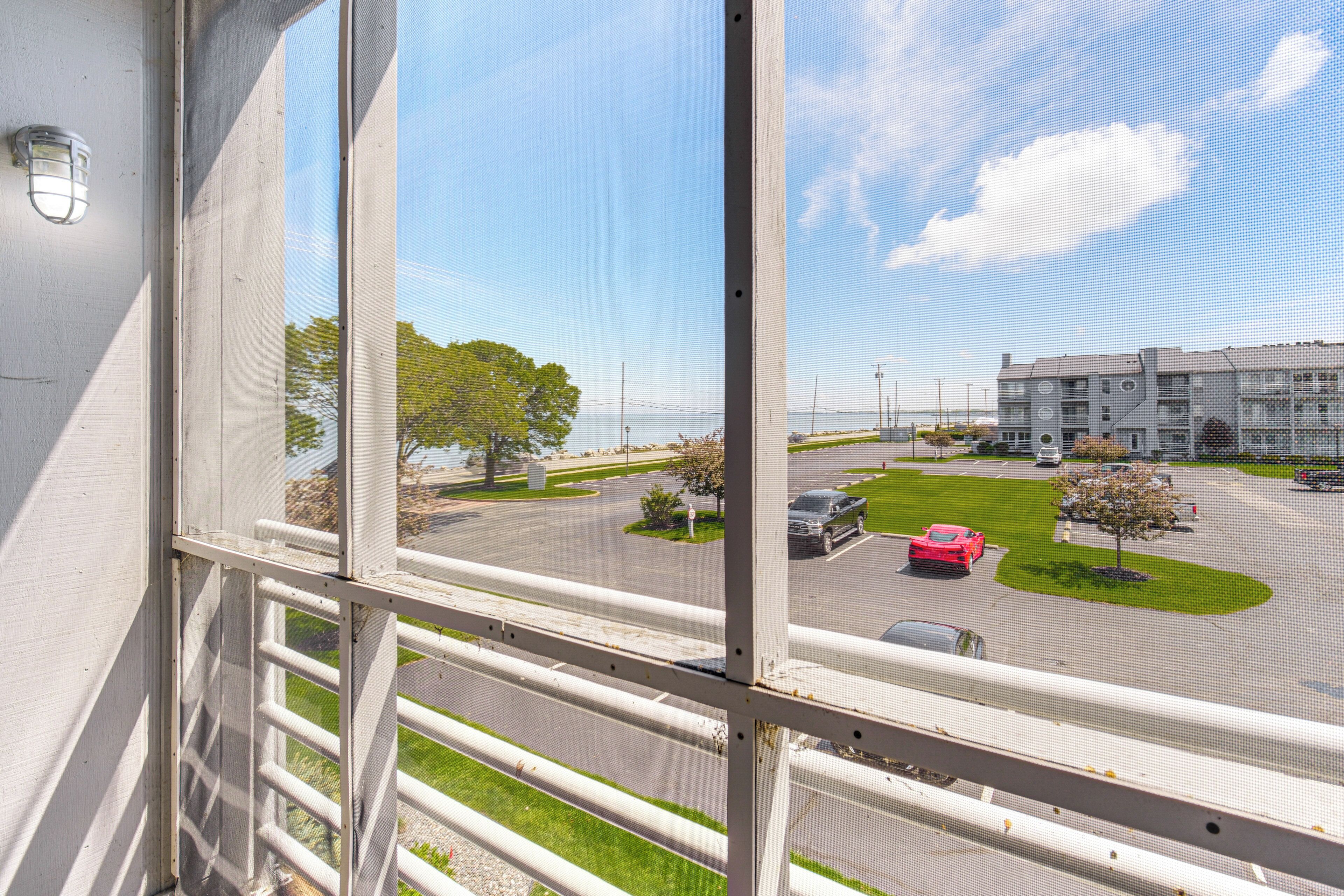 Blick durch ein Fenster mit Metallgitter auf einen Parkplatz mit einem roten Auto, ein mehrstöckiges Gebäude und einen klaren Himmel.