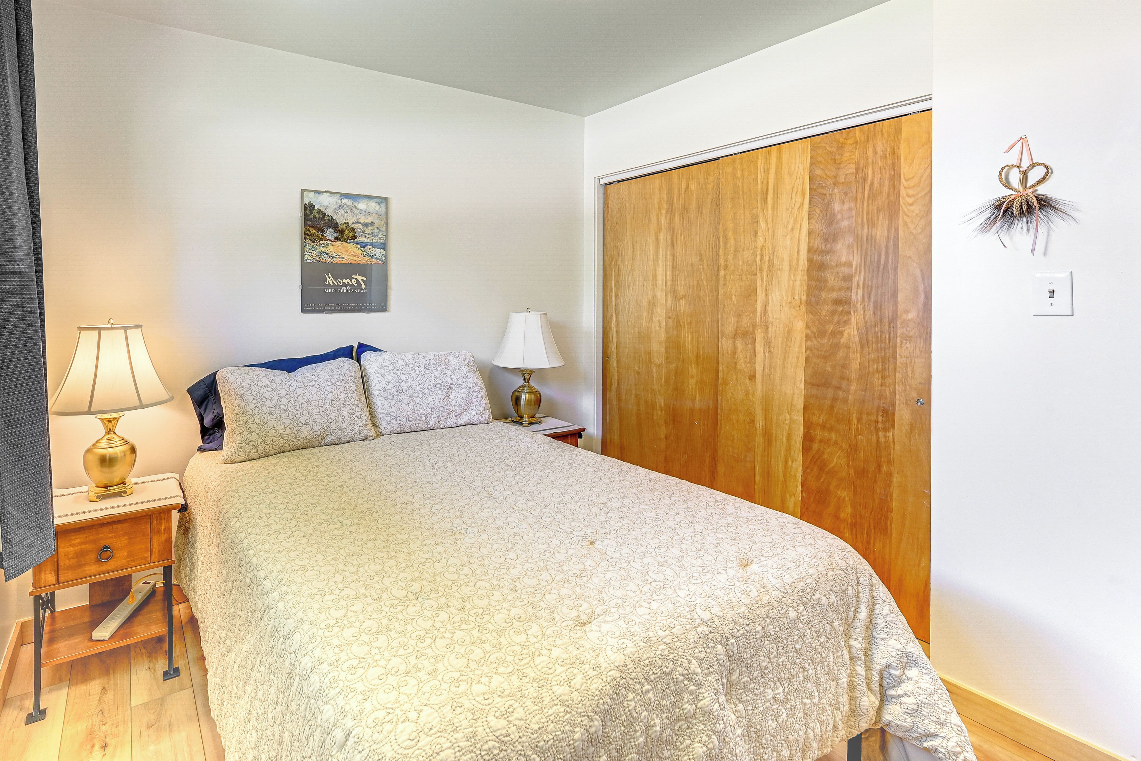 Una cama bien hecha con un edredón estampado, una mesita de noche con lámpara, un armario de madera y un cuadro colgado en la pared.