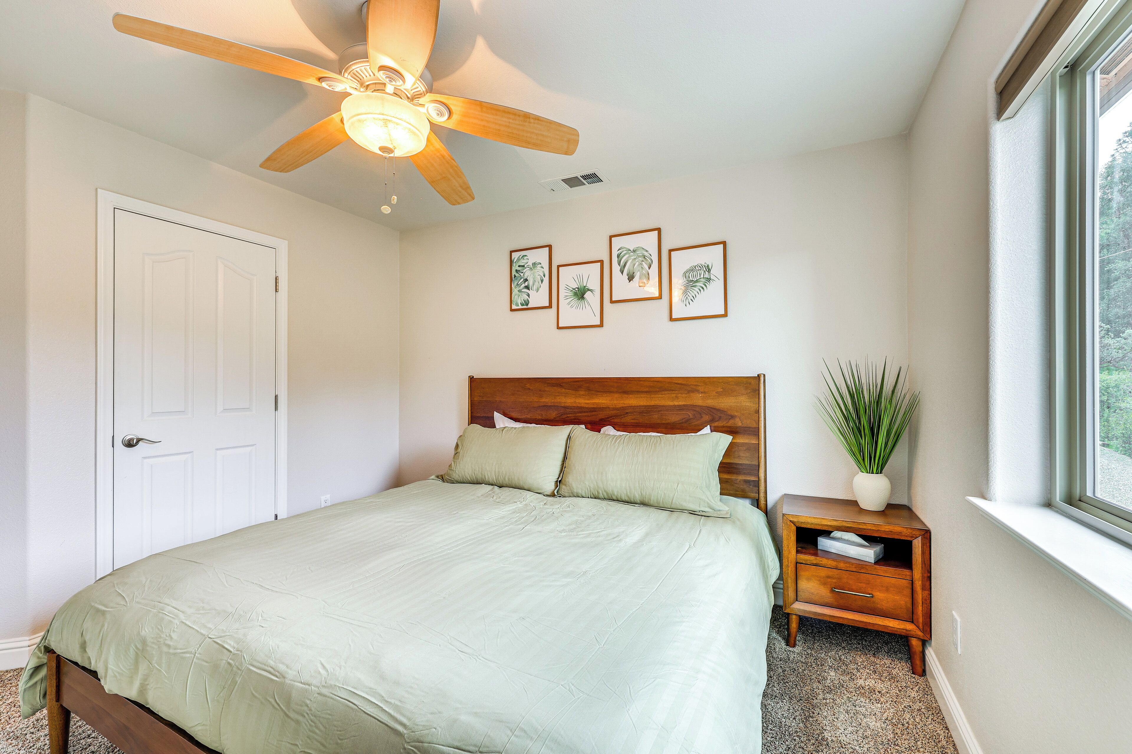 Une chambre à coucher avec un lit, un ventilateur de plafond, une table de chevet, une fenêtre et des tableaux encadrés accrochés au mur.