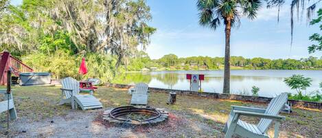 An outdoor seating area with Adirondack chairs, a fire pit, and a view of a lake surrounded by trees.