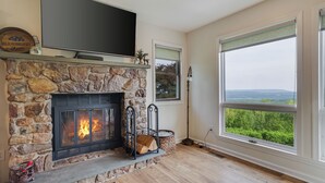 A stone fireplace with a TV above it, a large window with a view, and a basket of firewood. - Unreal Mountain Views w/ Hot tub Near Charming Jim Thorpe & Blue Mountain Skiing (Jim Thorpe)