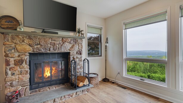 A stone fireplace with a TV above it, a large window with a view, and a basket of firewood.